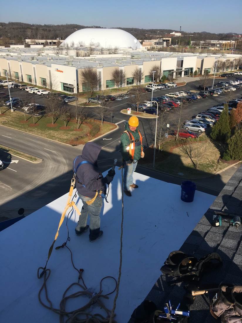 Wide view of a flat roof under construction on an apartment building in Knoxville, Tennessee, with workers on a safety line and the city skyline in the background
