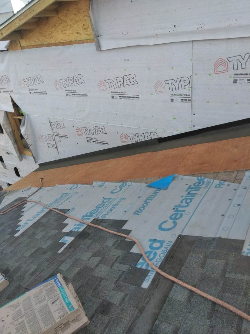 Synthetic roof underlayment meeting house wrap at a wall tie-in, with a bundle of shingles staged, new-construction apartment building near downtown Nashville, Tennessee